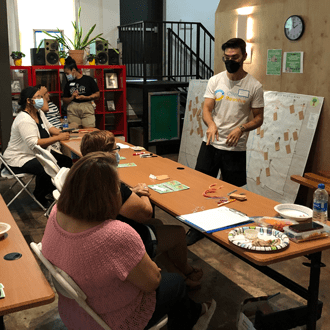 Facilitator leading a small group workshop in Boyle Heights, standing beside a presentation board while participants sit at tables with materials and engage in discussion