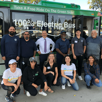 Group photo of people posing in front of a green electric bus with signage reading “100% Electric Bus” and “Take A Free Ride On The Green Side,” with participants standing and kneeling together outdoors