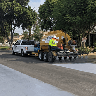 Worker applying cool pavement coating to a residential street using a spray truck, creating a lighter surface to reduce heat and improve urban climate resilience