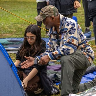 Instructor demonstrating emergency preparedness techniques to a participant setting up a tent outdoors, representing community resilience and disaster readiness