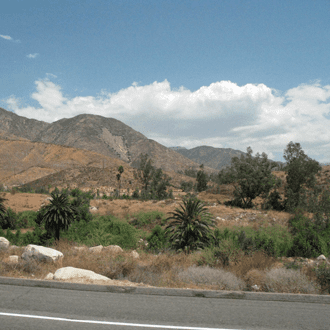 Landscape view of San Bernardino featuring dry hills and mountains in the background, with scattered trees and vegetation in the foreground under a partly cloudy sky