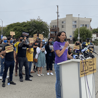 Woman speaking at a podium with microphones during an Our Future Los Angeles press event, with a group of supporters holding signs gathered behind her outdoors