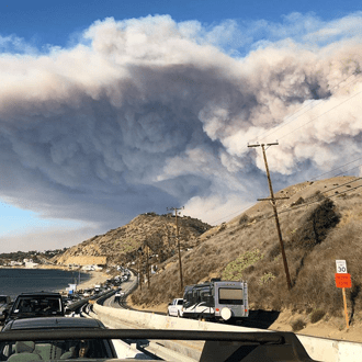 Climate Resolve Wildfire Smoke Over Highway Large plume of wildfire smoke rising over hills and a coastal highway with cars, illustrating climate-driven wildfire impacts and air quality concerns